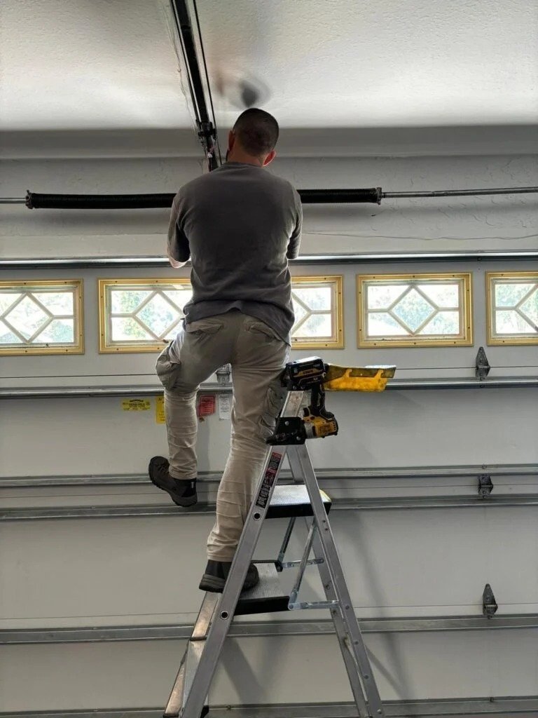 A person on a ladder holding a cordless drill beside a torsion spring